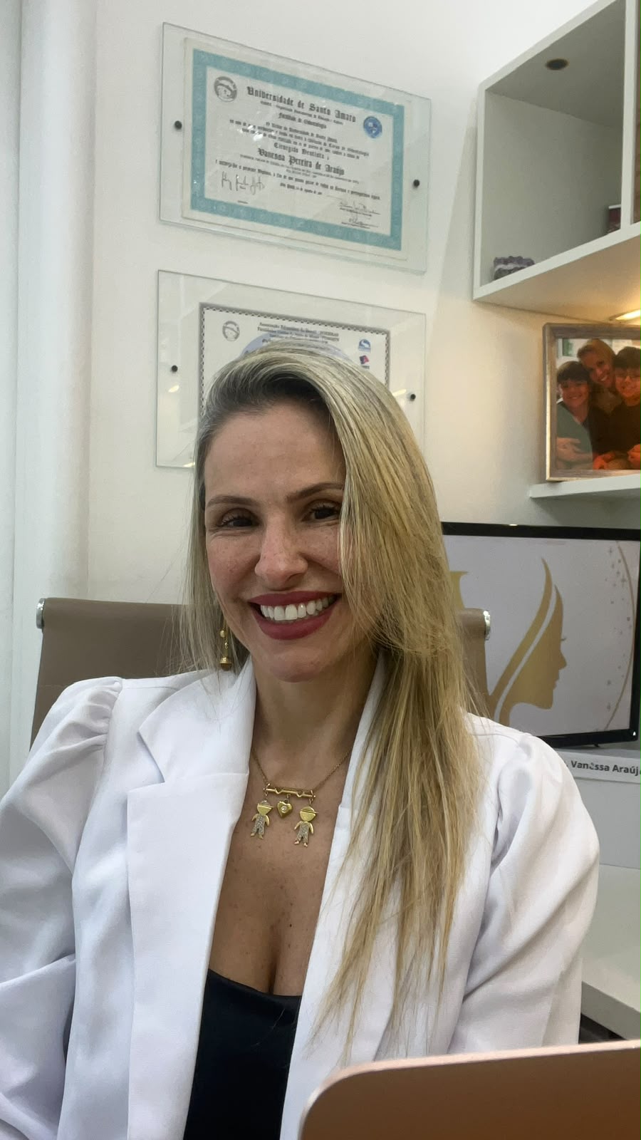 Dra. Vanessa Araújo de jaleco no consultório, com o diploma de Cirurgiã-Dentista da Universidade de Santo Amaro na parede